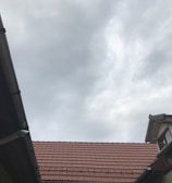 A view of rooftops with reddish-brown tiles, framed by wooden and metal guttering, and an overcast sky with grey clouds overhead.