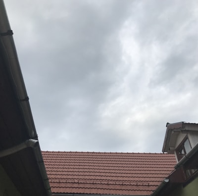 A view of rooftops with reddish-brown tiles, framed by wooden and metal guttering, and an overcast sky with grey clouds overhead.