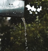 A metal gutter with water flowing from it in a smooth stream, surrounded by droplets. The background consists of blurred green foliage, suggesting a natural setting.