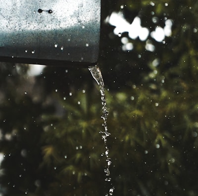 A metal gutter with water flowing from it in a smooth stream, surrounded by droplets. The background consists of blurred green foliage, suggesting a natural setting.