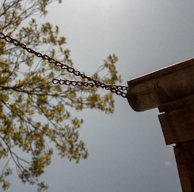 A metal gutter attached to a building with chains extends outward, with a tree branch in soft focus in the background against a blue sky. The sun casts a bright light, creating a serene and rustic atmosphere.