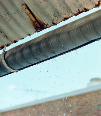 A metal gutter is attached to the edge of a corrugated roof, with water droplets dripping down. The background shows a blurred brick wall and a blue sky.