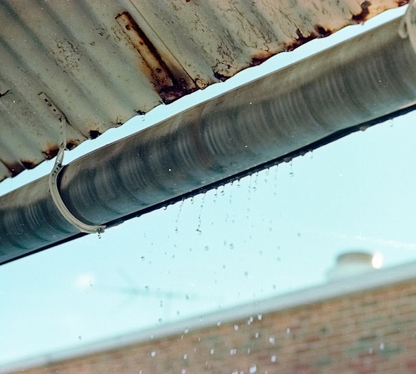 A metal gutter is attached to the edge of a corrugated roof, with water droplets dripping down. The background shows a blurred brick wall and a blue sky.