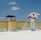 A person wearing protective gear is applying a white substance on a flat surface, possibly for insulation or coating. The individual is on a rooftop with a clear blue sky overhead. There are power lines in the background and a structure resembling a chimney.