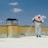 A person wearing protective gear is applying a white substance on a flat surface, possibly for insulation or coating. The individual is on a rooftop with a clear blue sky overhead. There are power lines in the background and a structure resembling a chimney.