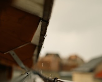 A close-up view of a rain gutter, capturing a droplet of water creating a splash as it hits the surface. The gutter is aligned with the edge of a roof, and the background appears blurred, highlighting the sharpness of the water droplet.