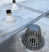 A rooftop surface with a metal drain cover and pipes protruding from the surface. The drain cover has vertical slats surrounding it. The surface appears to be slightly worn with some darker patches and dirt marks.