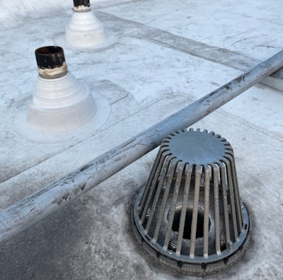 A rooftop surface with a metal drain cover and pipes protruding from the surface. The drain cover has vertical slats surrounding it. The surface appears to be slightly worn with some darker patches and dirt marks.